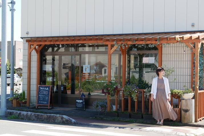 コーヒースタンド「Odaka Micro Stand Bar～オムスビ～」の前で風にあたる蒔田志保さんの写真。