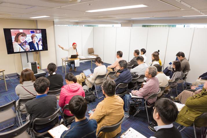 パーティションで仕切られた区画の中に設置された大型テレビを大勢の男女が視聴し、その横で小松さんがマイクを片手にテレビを指差しながら解説している写真