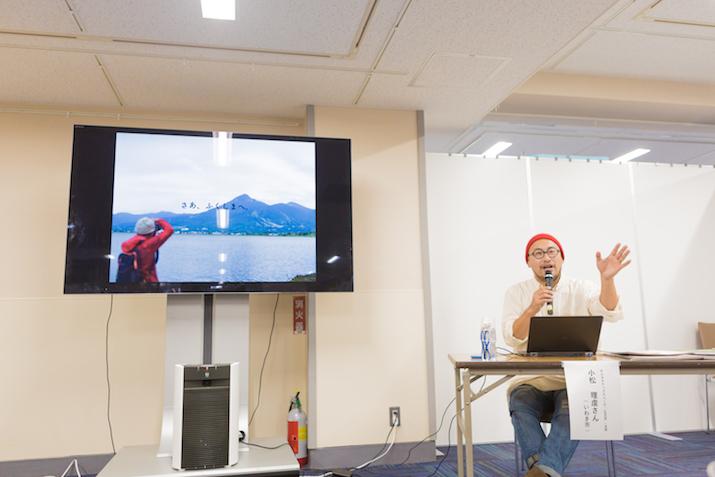 海の向こうに見える山を写真撮影する人とその上に「さあ、ふくしまへ」という字幕が表示された動画が流されているテレビと、その横でマイクを片手に解説する小松さんの写真